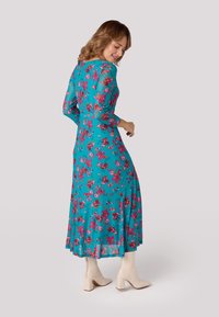 Langsleeve, teal jurk met een roze bloemenpatroon, met een aansluitend lijfje en een vloeiende rok. Gekleed met crèmekleurige enkellaarsjes.