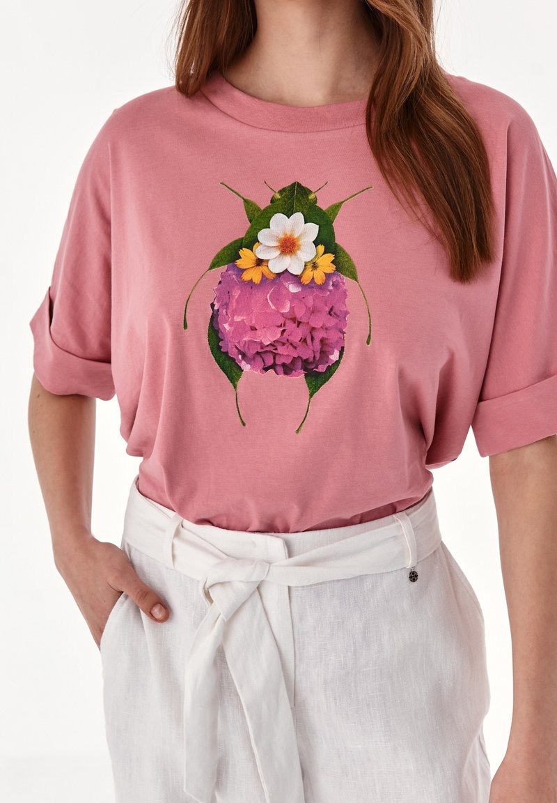 Femme portant une chemise rose avec un motif coloré de scarabée fleuri et un pantalon blanc taille haute avec une ceinture nouée.