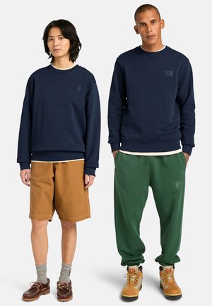 Zwei Models stehen nebeneinander und tragen marineblaue Sweatshirts; das eine kombiniert mit braunen Shorts und Bootsschuhen, das andere mit grünen Jogginghosen und Wanderschuhen.