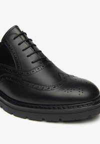 Scarpa brogue in pelle nera con perforazioni dettagliate, chiusura con lacci e suola in gomma. Presenta una finitura lucida e raffinata con cuciture a contrasto.