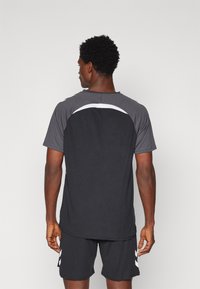 Zwarte sport t-shirt met grijze schouders en een witte accent bij de hals. Korte mouwen, gladde textuur en een relaxte pasvorm. Gecombineerd met zwarte shorts.