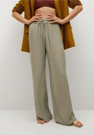 Femme portant un pantalon ample gris clair à taille ajustable, associé à une veste moutarde, un crop top bordeaux et des sandales beiges ouvertes.