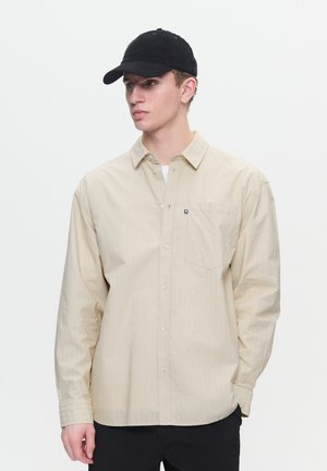 Beige long-sleeve shirt with vertical stripes, buttoned front, single chest pocket; worn with a black cap. Smooth, lightweight fabric.