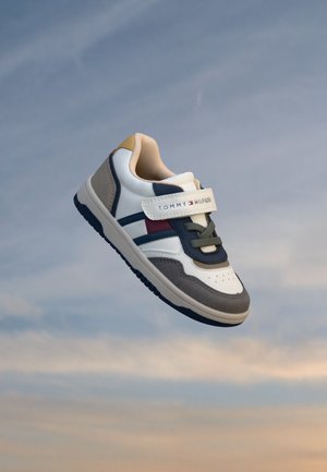 Baskets pour enfants en blanc, marine et beige, avec une bride à logo Tommy Hilfiger, flottant sur un fond de ciel doux et nuageux.