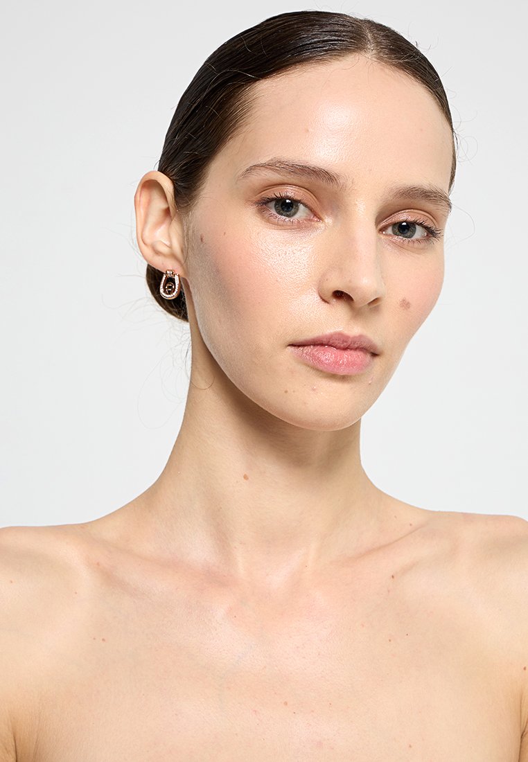 Goldene Creolen mit strukturierter Oberfläche, die ein zentrales rundes Design und kleine Metallakzente aufweisen. Das Model hat glatte Haut und einen neutralen Hintergrund.