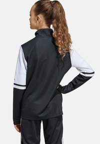 Schwarze und weiße sportliche Jacke mit hohem Kragen, langen Ärmeln und strukturiertem Stoff mit kontrastierenden Streifen an den Ärmeln.