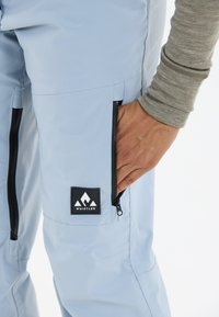 Ljusblåa vattentäta byxor med sidofickor med blixtlås och en svart logotypbricka med texten "WHISTLER." Tyget har en slät, strukturerad yta.