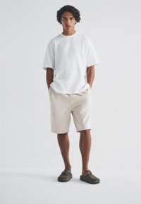 Weißes Kurzarmhemd, beige Shorts und braune Clogs. Lässige Passform, weicher Stoff und entspannter Schnitt. Einfaches Design ohne Muster oder Logos.