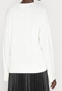 Vit stickad tröja med avslappnad passform, med rund halsringning och ribbade ärmslut, tillsammans med en svart plisserad kjol.