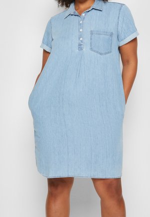 Kobieta ubrana w jasnoniebieską sukienkę jeansową z krótkim rękawem, kołnierzykiem, plisą na guziki, kieszonką na piersi i dłońmi w bocznych kieszeniach.