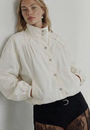 Blondynka ubrana w białą kurtkę zapinaną pod szyję, z rękami w kieszeniach oraz czarną koronkową spódnicę z paskiem, stojąca na tle białej ściany.