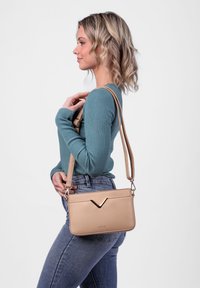 Hnědá crossbody taška s hladkou texturou, V-článkovým akcentem na přední straně a nastavitelným popruhem. Jednoduchý design ideální pro každodenní použití.