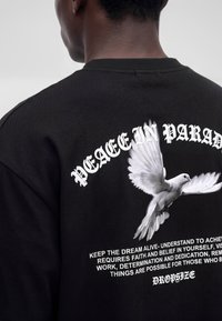 Person wearing a black shirt with a white flying dove and "PEACE IN PARADISE" text above in gothic font on the back.