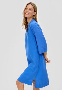 Une robe en lin bleu présente des manches trois-quarts, un ourlet fendu et une silhouette décontractée. Le tissu semble léger et texturé.