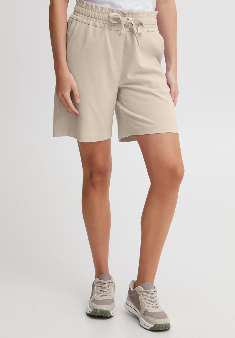 Shorts de sport beiges avec une taille élastique et un cordon de serrage. Fabriqués en tissu lisse, avec des poches latérales. Une paire de baskets grises et blanches en dessous.