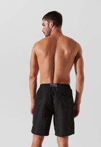 Schwarze Badeshorts mit elastischem Bund und Seitentasche. Glatte Textur, knielanges Design und dezente Markenkennzeichnung auf der Rückseite.