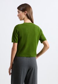 Zielony sweter z krótkim rękawem z miękkiego materiału, dopasowany krój, okrągły dekolt i prążkowane wykończenia, zestawiony z szarymi spodniami z wysokim stanem.