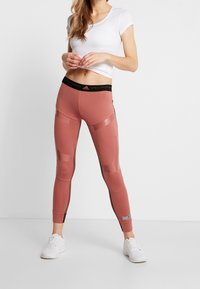 Rosa leggings med en hög svart midjeresår, försedda med glossy partier och en tajt passform. Kombinerade med en vit topp med korta ärmar och vita sneakers.