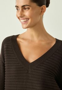 Kvinde smilende, hovedet drejet til højre, iført en mørkebrun ribstrikket V-hals sweater og små guldringe, med håret trukket tilbage.