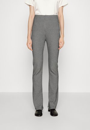 Femme portant un pantalon slim taille haute à motif pied-de-poule noir et blanc, associé à des chaussures noires et un top blanc à manches courtes.
