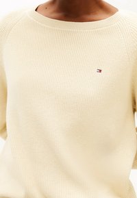 Pull en tricot couleur crème avec une texture côtelée, un col large et un petit logo rouge, blanc et bleu sur la poitrine.