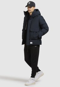 Marineblå pufferjakke med hætte, som har to forlommer. Kombineret med sorte sweatpants og hvide high-top sneakers.