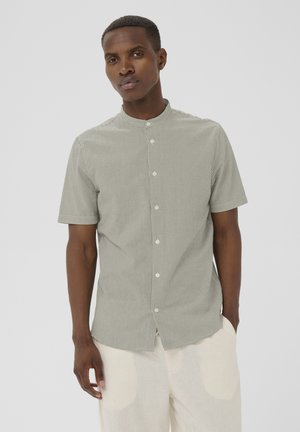 Man draagt een lichtgroen slim-fit overhemd met korte mouwen, dunne verticale strepen, en beige broek, staand met één hand in de zak.