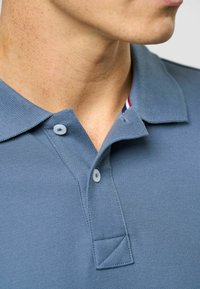 Zils polo krekls, kas izgatavots no teksturētas auduma, ar apkakli, divām gaišām pogām un svītrainu detaļu zem apkakles.