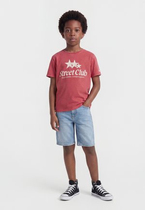 Garçon debout, main dans la poche, portant un t-shirt rouge "Street Club", un short en jean clair et des baskets montantes noires sur fond blanc.