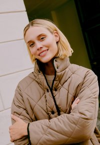 Quilted tan jacket with black trim and buttons, worn over a neutral sweater. The model has short blonde hair and is standing outdoors.
