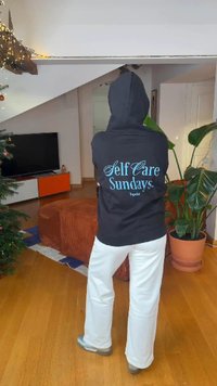 Sweat à capuche noir avec l'inscription "Self Care Sundays." en bleu clair. Pantalon blanc à jambes larges. Cadre intérieur avec une plante et des meubles.