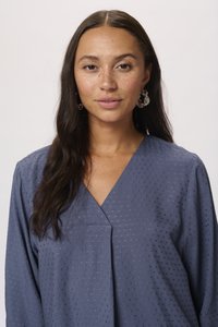 Blusa blu, strutturata, con scollatura a V e motivo a pois. Maniche lunghe, tessuto morbido e vestibilità dritta esaltano il suo design. Orecchini d'argento visibili.