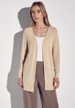 Cardigan beige à manches longues avec un design à l'avant ouvert, fabriqué en tissu texturé. Assorti avec un débardeur blanc et un pantalon marron.
