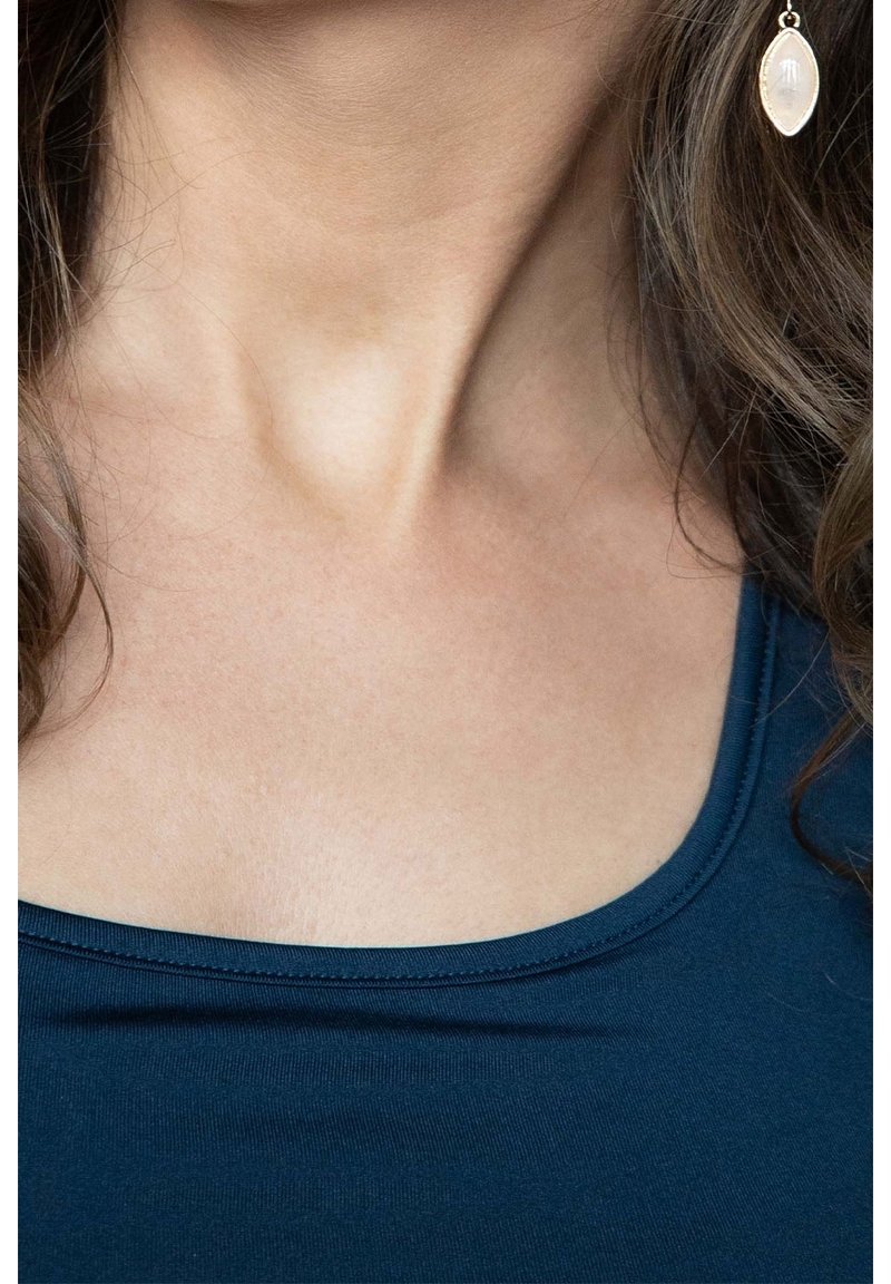 Gros plan sur le cou et le haut de la poitrine d’une femme portant un haut bleu foncé et une boucle d’oreille délicate avec un pendentif translucide.
