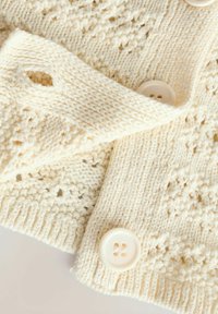 Gros plan d’un tissu tricoté couleur crème avec des motifs détaillés et deux boutons ronds crème attachés par des boutonnières.