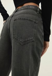 Pantalones vaqueros de mezclilla negra con corte recto, que presentan un bolsillo trasero y un acabado texturizado. La cinturilla cuenta con una etiqueta en forma de rectángulo.