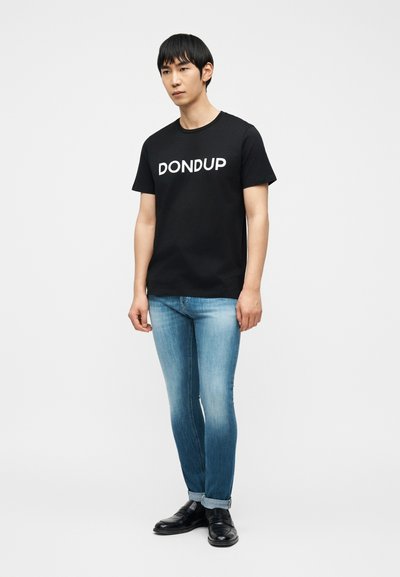 Νέος άνδρας όρθιος, φορώντας μαύρο t-shirt "DONDUP", ξεθωριασμένο μπλε τζιν με γυρισμένα μανίκια και μαύρα δερμάτινα επίσημα παπούτσια σε λευκό φόντο.