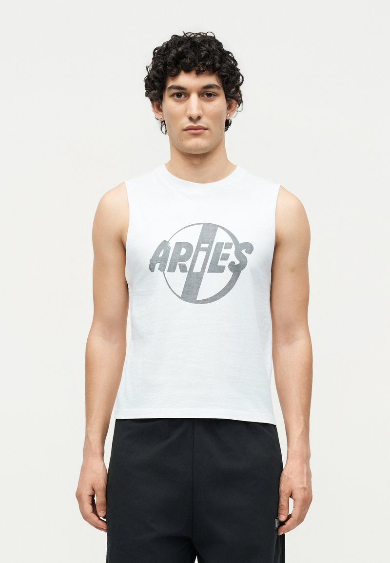 Joven con pelo rizado que lleva una camisa blanca sin mangas con el logo "ARIES" y pantalones negros, posando contra un fondo claro y liso.