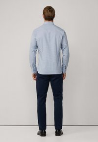 Muž stojaci chrbtom, oblečený v svetlomodrej košeli s dlhým rukávom, námorníckych nohaviciach a čiernych spoločenských topánkach na jednotnom pozadí.