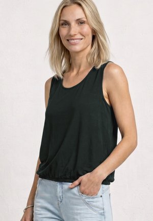 Smilende kvinde med blondt hår iført en ærmeløs sort top og lyseblå jeans, stående med den ene hånd i lommen.