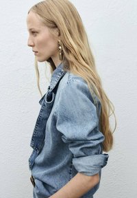 Profil d'une femme aux longs cheveux blonds portant une veste en denim retroussée et de grandes boucles d'oreilles en or, sur fond blanc uni.