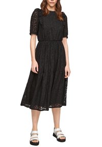 Robe noire avec un motif floral texturé, des manches courtes bouffantes, une taille élastique et une jupe à volants. Portée avec des sandales blanches.
