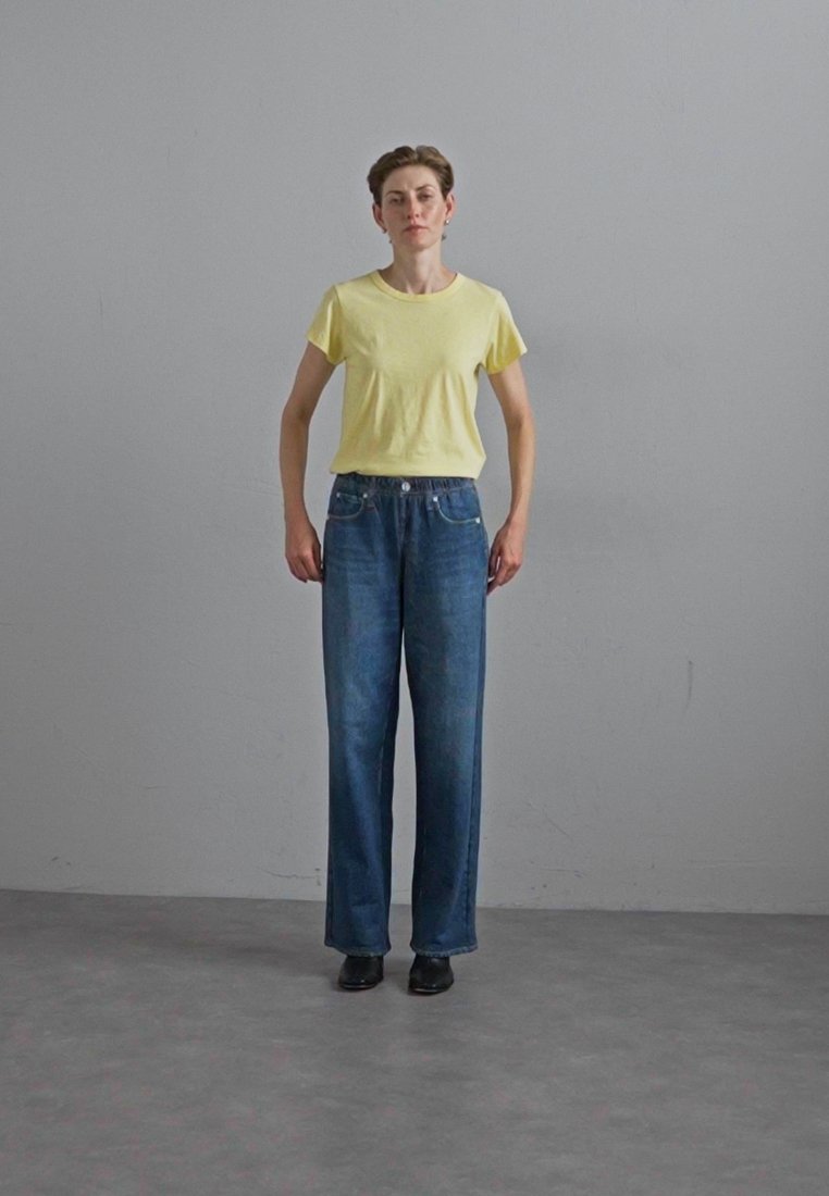 T-shirt en coton jaune, coupe ample, associé à un jean bleu en denim taille haute, design à jambe droite, avec peu de détails, porté avec des chaussures noires.
