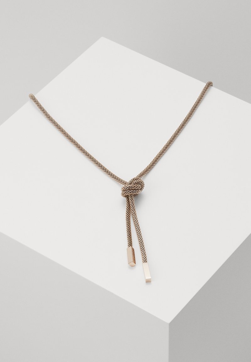 Collier fin en cordon tressé beige et marron noué en un simple nœud avec deux embouts métalliques rectangulaires sur un socle blanc.