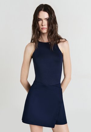 Junge Frau mit langen, welligen Haaren trägt ein ärmelloses, dunkelblaues Kleid mit einem asymmetrischen Rock vor einem schlichten, hellen Hintergrund.