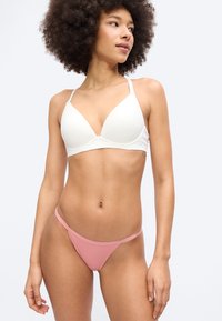 Γυναίκα με σγουρά μαλλιά που φοράει λευκό σουτιέν και ροζ εσώρουχα, στέκεται με το κεφάλι γυρισμένο προς τα δεξιά σε απλό φωτεινό φόντο.