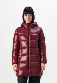 Vrouw in een glanzende bordeauxrode gewatteerde pufferjas met capuchon, ritssluiting aan de voorkant, zijzakken en een zwarte ronde patch op de linkermouw.