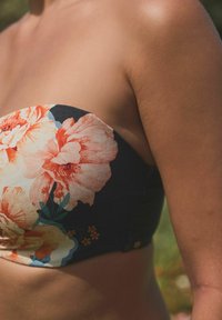 Bikini-topp utan axelband med en mörk bas och stora, färgglada blommönster i orange och rosa. Släta tyger med minimala metalldetaljer.