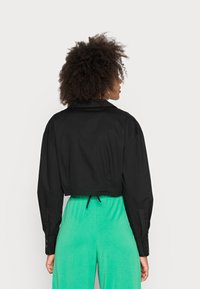 Casaco curto preto com colarinho estruturado, mangas longas e punhos abotoados, usado sobre um conjunto verde, apresentando um cordão na bainha.