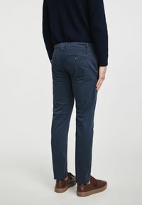 Osoba stoi tyłem, ubrana w granatowe spodnie i brązowe trampki, w ciemnym swetrze, prezentując widok pleców swojej stylizacji.